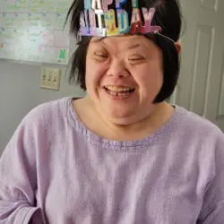 A woman wearing a multicolor Happy Birthday crown and purple shirt is shown closeup with a big smile. Behind her a white interior door and a white board calendar are visible along with a gray wall.