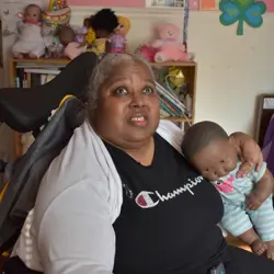 A woman is sitting in a wheelchair, holding a doll in her left arm. She is wearing a black 'Champion' shirt and a light gray cardigan. Behind her, there is a bookshelf with several dolls and stuffed animals. A bed with a purple and white blanket is visible on the right side of the image.