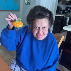An older woman is seated indoors, holding up a small, orange pumpkin decoration with a green bow. She is wearing a bright blue sweater and checkered pants. The background shows a kitchen area with dark cabinets and a blue bulletin board with decorations.