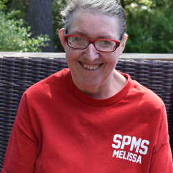 A white woman with short hair and wearing a bright red shirt is seated and smiling outdoors.