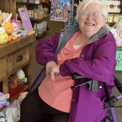 An older woman with short white hair and glasses is smiling warmly while sitting in a purple walker seat. She is wearing a coral-orange top and a purple jacket, and is posed in a store filled with Easter-themed stuffed animals and decorations. A wooden display table to her left showcases plush bunnies and toys, and colorful signs and holiday items are visible on shelves behind her. The setting is bright and cheerful, suggesting a festive shopping outing.