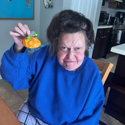 An older woman is seated indoors, holding up a small, orange pumpkin decoration with a green bow. She is wearing a bright blue sweater and checkered pants. The background shows a kitchen area with dark cabinets and a blue bulletin board with decorations.